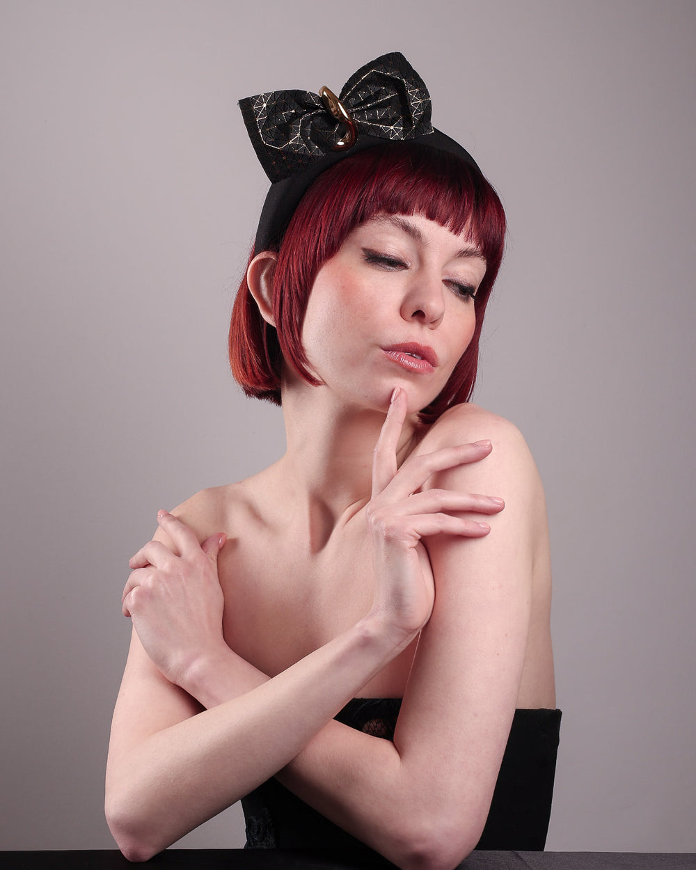 The model is wearing and showcasing the right front below angle of the Valkyrie, which features a sculpted bow headband with a gold ring detail. This holds a structured bow in black faux leather with a gold metallic base. The bow is dramatically placed across a plush headband base cinched by the gold-tone ring. Designed by Merve Bayindir.