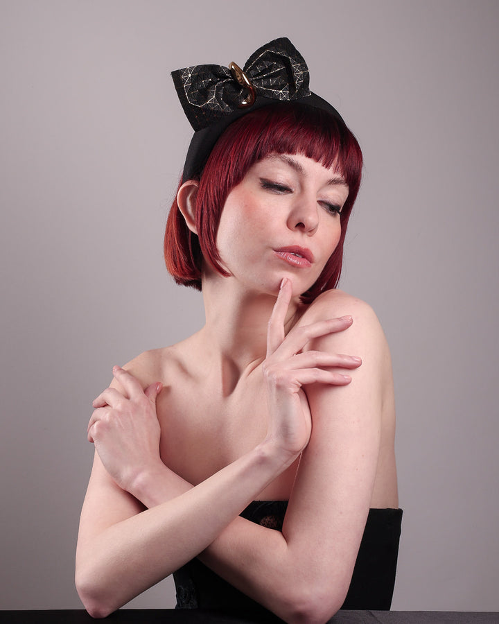 The model is wearing and showcasing the right front below angle of the Valkyrie, which features a sculpted bow headband with a gold ring detail. This holds a structured bow in black faux leather with a gold metallic base. The bow is dramatically placed across a plush headband base cinched by the gold-tone ring. Designed by Merve Bayindir.
