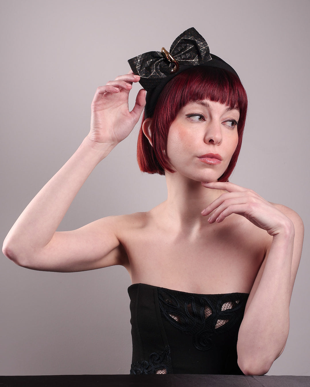 The model is wearing and showcasing the right front of the Valkyrie, which features a sculpted bow headband with a gold ring detail. This holds a structured bow in black faux leather with a gold metallic base. The bow is dramatically placed across a plush headband base cinched by the gold-tone ring. Designed by Merve Bayindir.