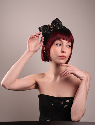 The model is wearing and showcasing the right front of the Valkyrie, which features a sculpted bow headband with a gold ring detail. This holds a structured bow in black faux leather with a gold metallic base. The bow is dramatically placed across a plush headband base cinched by the gold-tone ring. Designed by Merve Bayindir.