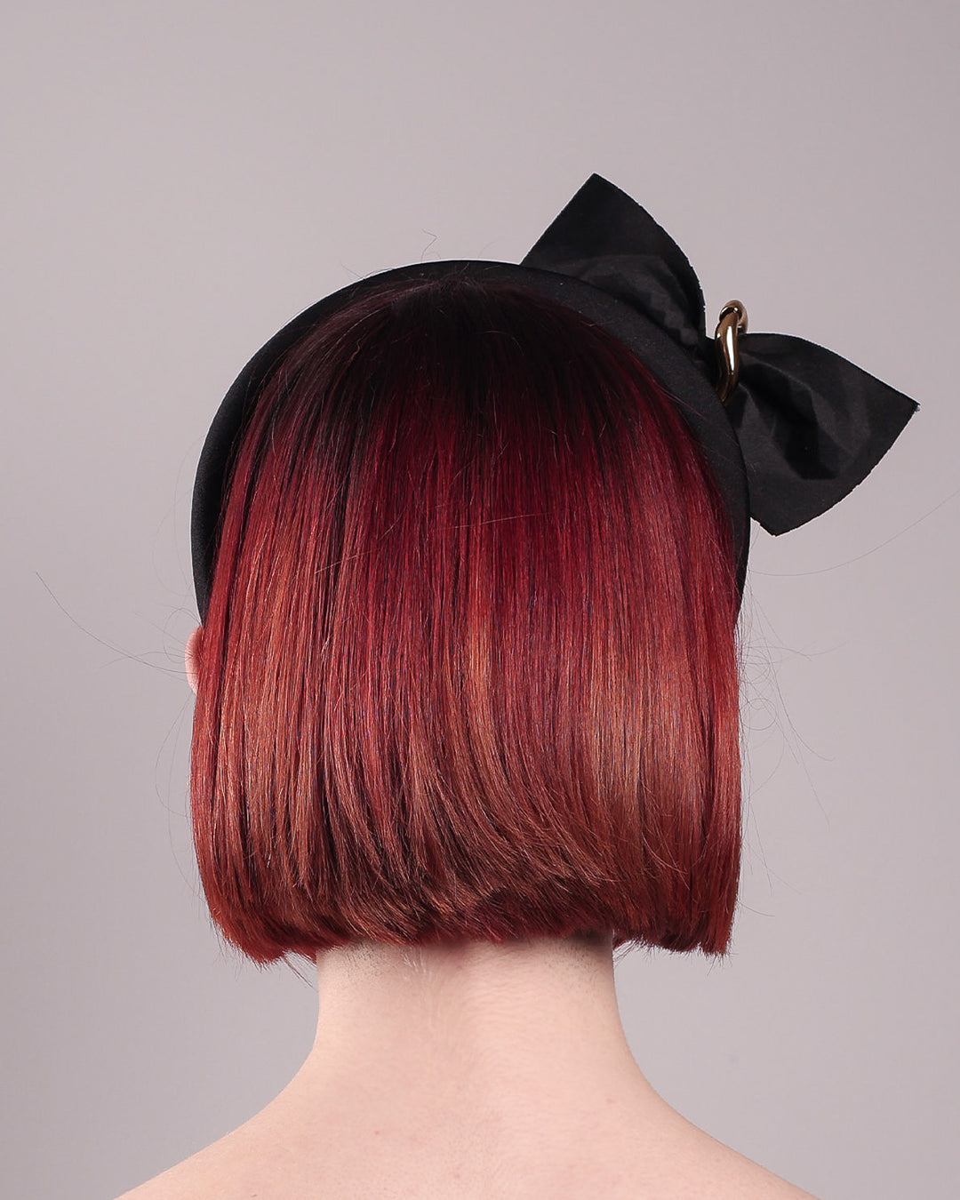 The model is showcasing the back of the Valkyrie, which features a sculpted bow headband with a gold ring detail. This holds a structured bow in black faux leather with a gold metallic base. The bow is dramatically placed across a plush headband base cinched by the gold-tone ring. Designed by Merve Bayindir.