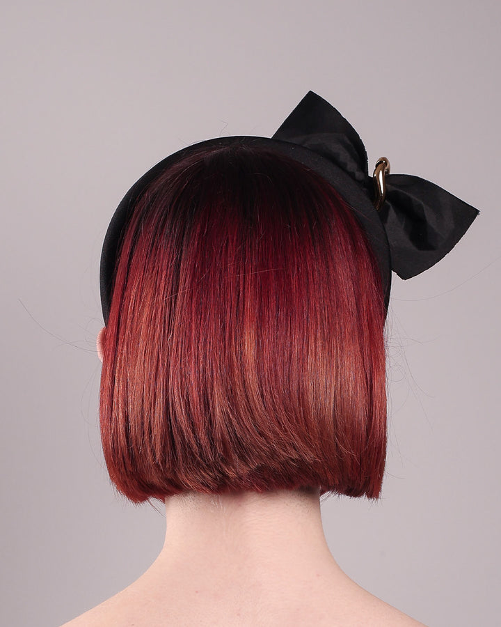 The model is showcasing the back of the Valkyrie, which features a sculpted bow headband with a gold ring detail. This holds a structured bow in black faux leather with a gold metallic base. The bow is dramatically placed across a plush headband base cinched by the gold-tone ring. Designed by Merve Bayindir.