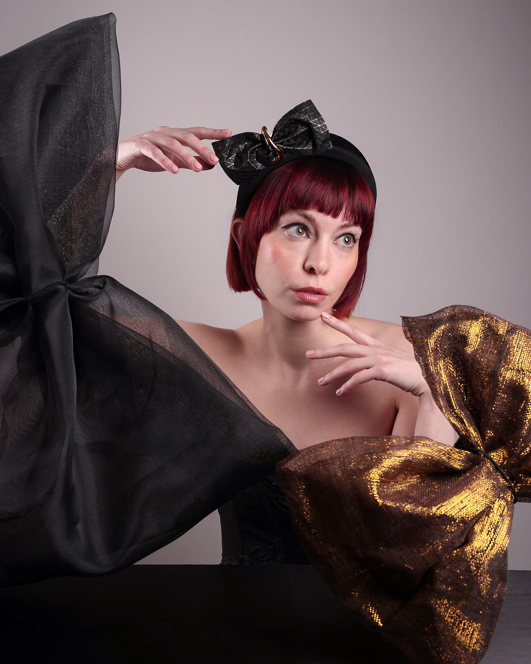 The editorial picture of the model posing with one black and one gold large bow decorations while showcasing the front of the Valkyrie, which features a sculpted bow headband with a gold ring detail. This holds a structured bow in black faux leather with a gold metallic base. The bow is dramatically placed across a plush headband base cinched by the gold-tone ring. Designed by Merve Bayindir.