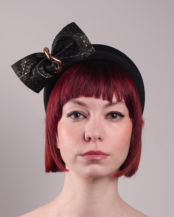 The model is showcasing the front close up of the Valkyrie, which features a sculpted bow headband with a gold ring detail. This holds a structured bow in black faux leather with a gold metallic base. The bow is dramatically placed across a plush headband base cinched by the gold-tone ring. Designed by Merve Bayindir.