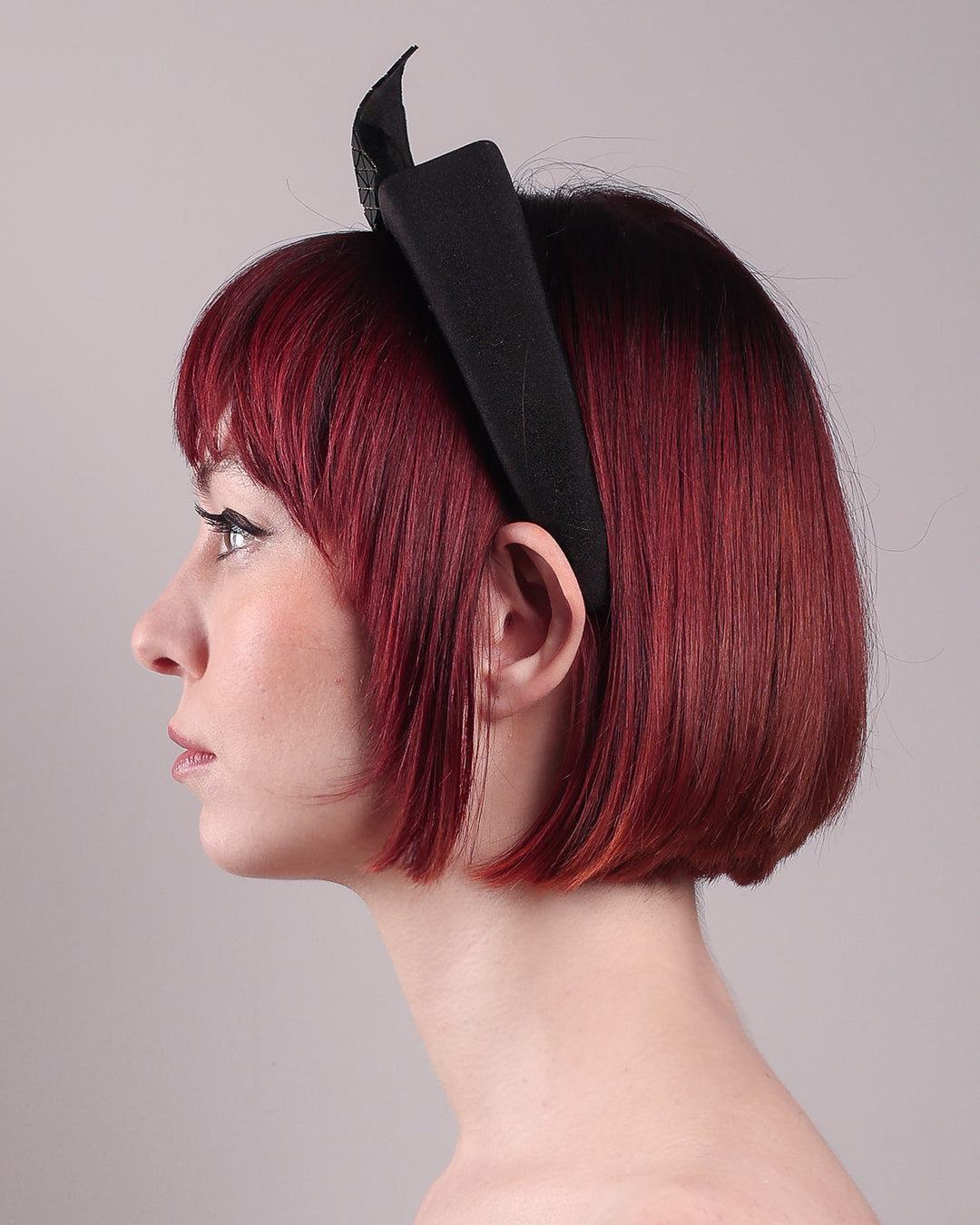 The model is showcasing the left close up of the Valkyrie, which features a sculpted bow headband with a gold ring detail. This holds a structured bow in black faux leather with a gold metallic base. The bow is dramatically placed across a plush headband base cinched by the gold-tone ring. Designed by Merve Bayindir.