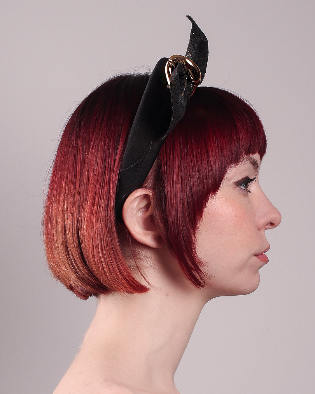 The model is showcasing the right close up of the Valkyrie, which features a sculpted bow headband with a gold ring detail. This holds a structured bow in black faux leather with a gold metallic base. The bow is dramatically placed across a plush headband base cinched by the gold-tone ring. Designed by Merve Bayindir.