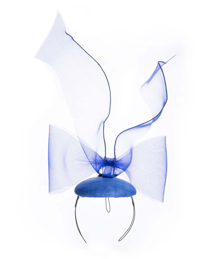 Still life picture of Royal blue button shaped fascinator decorated with a large crinoline bow and flying crinoline ribbons.