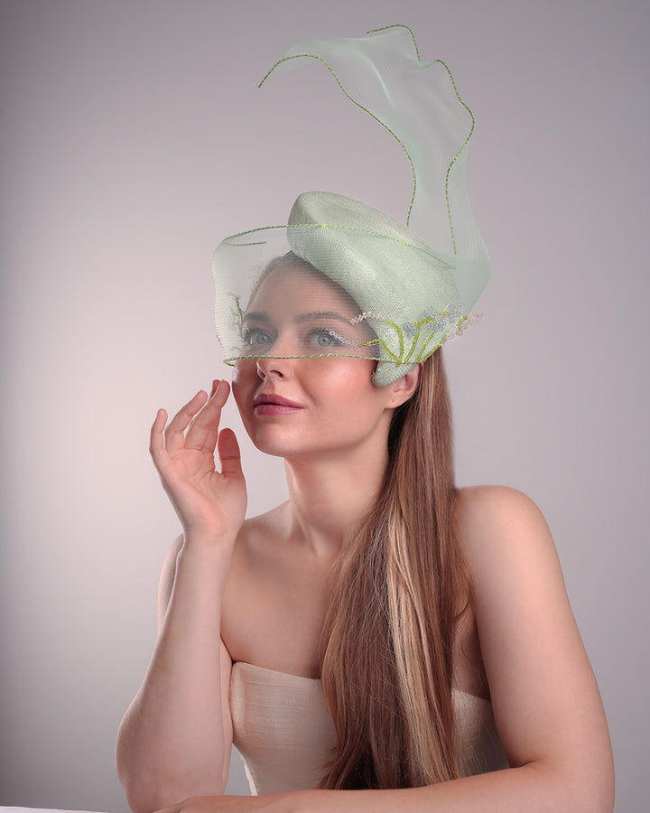 Model is showcasing the left front side of light grean tear drop fascinator moving from the top right of the face to the left cheek. The beautiful fascinator adorned with a scultured crinoline that covers the eyes in veil llike position and fly from side to back high creating a wind flown shawl effect. Both sides of the crinoline piece is decorated with bead embroidery to give it a toucsh of softness.