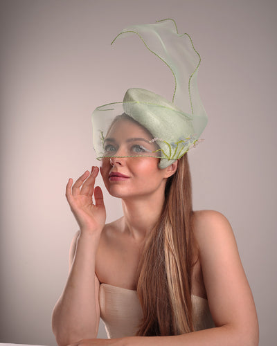 Model is showcasing the left front side of light grean tear drop fascinator moving from the top right of the face to the left cheek. The beautiful fascinator adorned with a scultured crinoline that covers the eyes in veil llike position and fly from side to back high creating a wind flown shawl effect. Both sides of the crinoline piece is decorated with bead embroidery to give it a toucsh of softness.