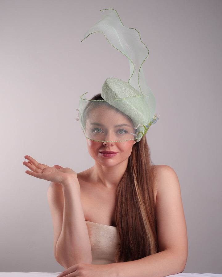 Model is showcasing thefront of light grean tear drop fascinator moving from the top right of the face to the left cheek. The beautiful fascinator adorned with a scultured crinoline that covers the eyes in veil llike position and fly from side to back high creating a wind flown shawl effect. Both sides of the crinoline piece is decorated with bead embroidery to give it a toucsh of softness.