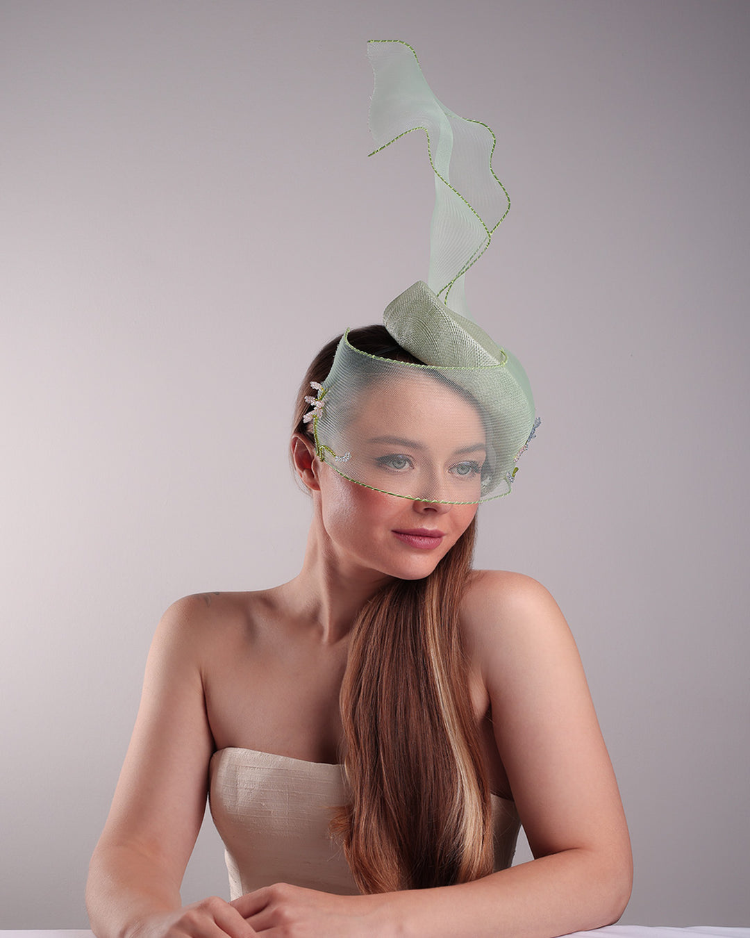 Model is showcasing the right front side of light grean tear drop fascinator moving from the top right of the face to the left cheek. The beautiful fascinator adorned with a scultured crinoline that covers the eyes in veil llike position and fly from side to back high creating a wind flown shawl effect. Both sides of the crinoline piece is decorated with bead embroidery to give it a toucsh of softness.