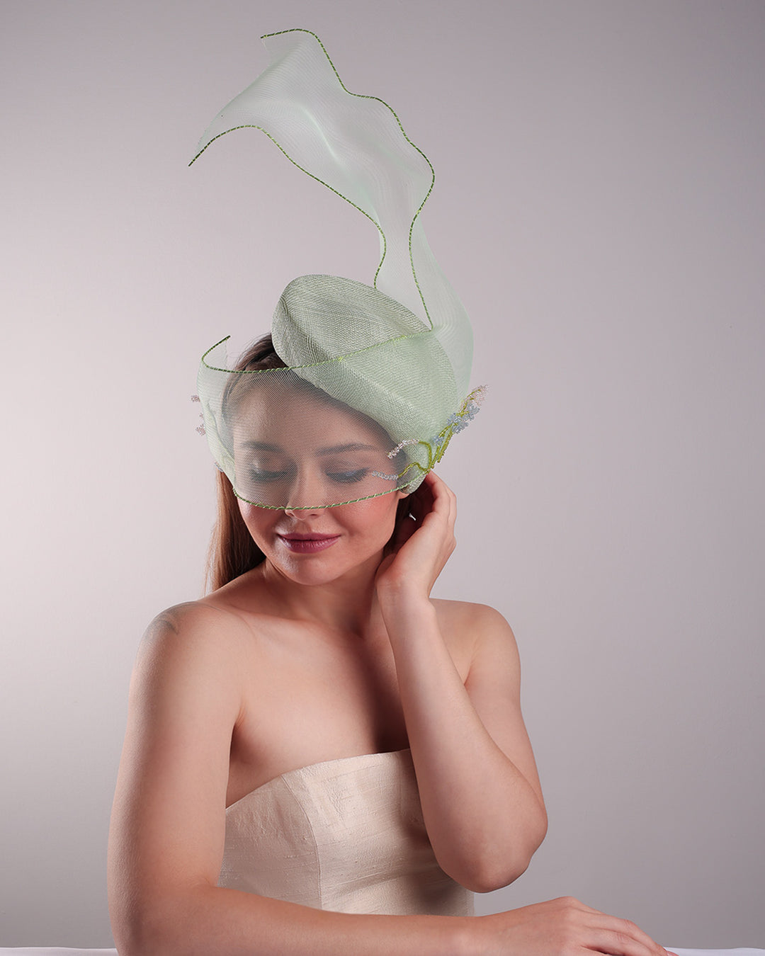 Model is showcasing the front abve of light grean tear drop fascinator moving from the top right of the face to the left cheek. The beautiful fascinator adorned with a scultured crinoline that covers the eyes in veil llike position and fly from side to back high creating a wind flown shawl effect. Both sides of the crinoline piece is decorated with bead embroidery to give it a toucsh of softness.