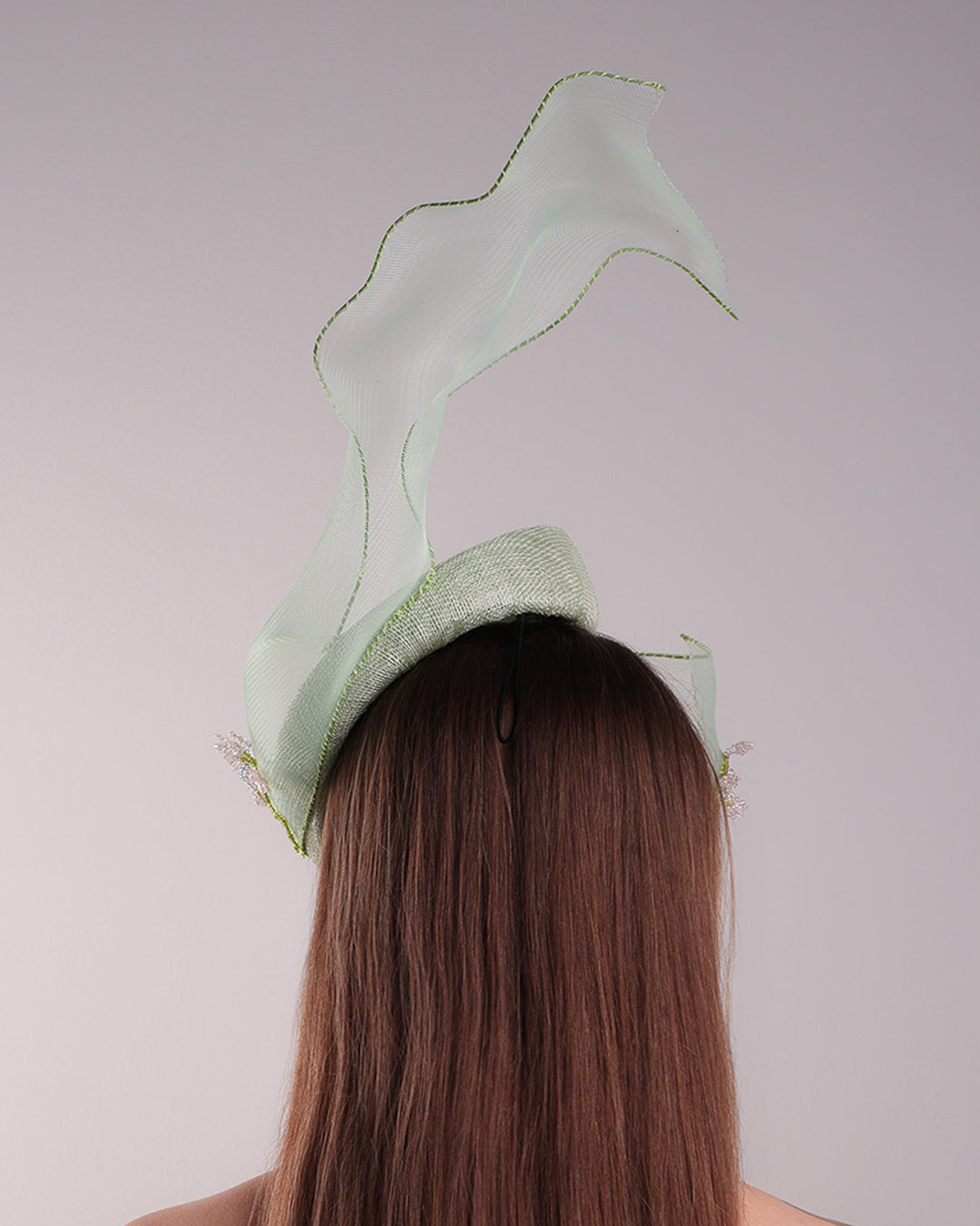 Model is showcasing the back of light grean tear drop fascinator moving from the top right of the face to the left cheek. The beautiful fascinator adorned with a scultured crinoline that covers the eyes in veil llike position and fly from side to back high creating a wind flown shawl effect. Both sides of the crinoline piece is decorated with bead embroidery to give it a toucsh of softness.