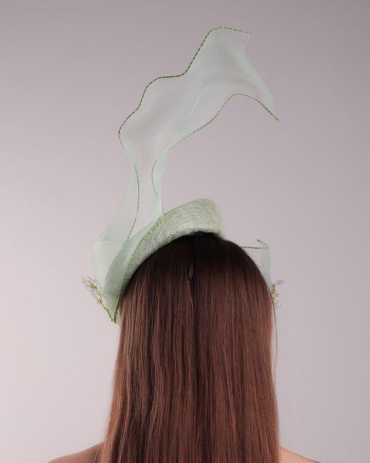Model is showcasing the back of light grean tear drop fascinator moving from the top right of the face to the left cheek. The beautiful fascinator adorned with a scultured crinoline that covers the eyes in veil llike position and fly from side to back high creating a wind flown shawl effect. Both sides of the crinoline piece is decorated with bead embroidery to give it a toucsh of softness.