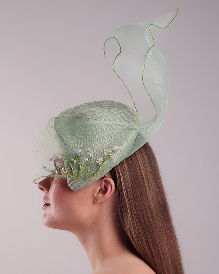 Model is showcasing the left side of light grean tear drop fascinator moving from the top right of the face to the left cheek. The beautiful fascinator adorned with a scultured crinoline that covers the eyes in veil llike position and fly from side to back high creating a wind flown shawl effect. Both sides of the crinoline piece is decorated with bead embroidery to give it a toucsh of softness.