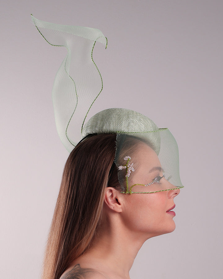 Model is showcasing the right side of light grean tear drop fascinator moving from the top right of the face to the left cheek. The beautiful fascinator adorned with a scultured crinoline that covers the eyes in veil llike position and fly from side to back high creating a wind flown shawl effect. Both sides of the crinoline piece is decorated with bead embroidery to give it a toucsh of softness.