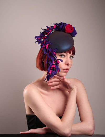 The model is showcasing a right front view of a pillbox fascinator with an ombré finish that transitions from deep midnight to luminous violet under varying light conditions. Adorned with handcrafted violet and fuchsia flowers, they cascade asymmetrically from the back to the right side of the face, extending to under the chin. The pillbox is fitted with a sleek headband and an elastic. Designed by Merve Bayindir.