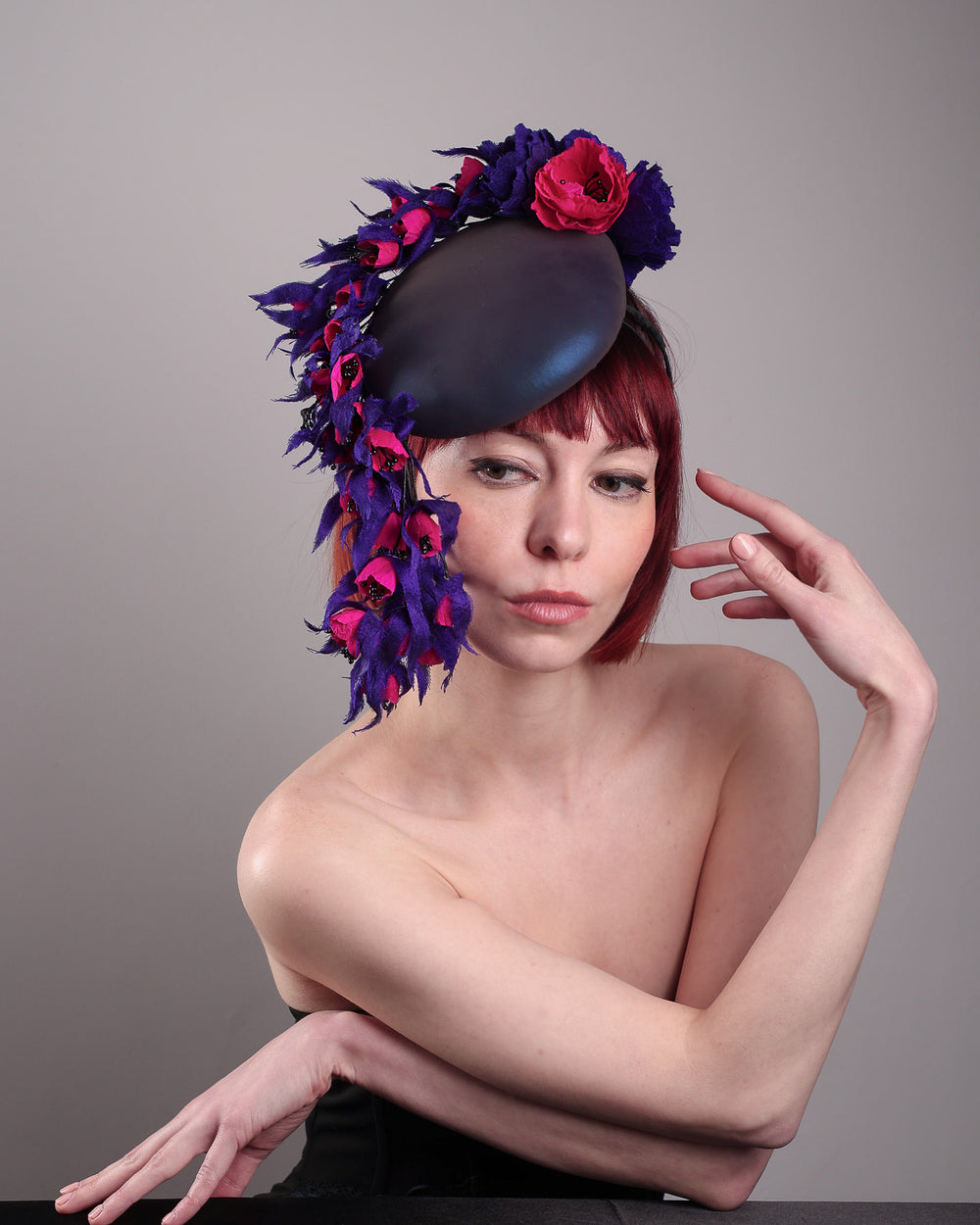 The model is showcasing the front view of a pillbox fascinator with an ombré finish that transitions from deep midnight to luminous violet under varying light conditions. Adorned with handcrafted violet and fuchsia flowers, they cascade asymmetrically from the back to the right side of the face, extending to under the chin. The pillbox is fitted with a sleek headband and an elastic. Designed by Merve Bayindir.