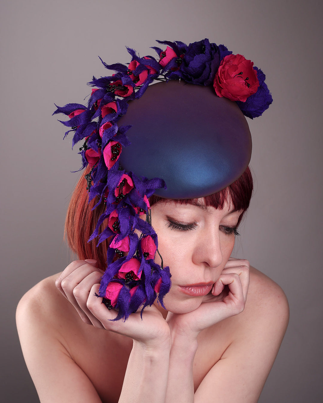 The model is showcasing the topt front view of a pillbox fascinator with an ombré finish that transitions from deep midnight to luminous violet under varying light conditions. Adorned with handcrafted violet and fuchsia flowers, they cascade asymmetrically from the back to the right side of the face, extending to under the chin. The pillbox is fitted with a sleek headband and an elastic. Designed by Merve Bayindir.