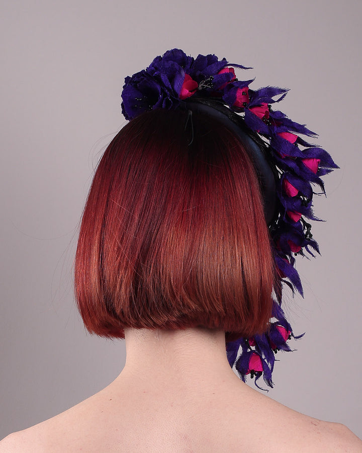 The model is showing the back view of a pillbox fascinator with an ombré finish that transitions from deep midnight to luminous violet under varying light conditions, on her head. Adorned with handcrafted violet and fuchsia flowers, they cascade asymmetrically from the back to the right side of the face, extending to under the chin. The pillbox is fitted with a sleek headband and an elastic. Designed by Merve Bayindir.