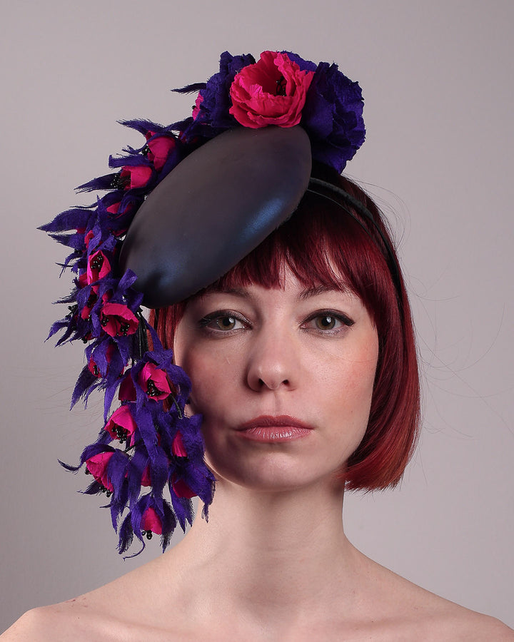 The model is showing the front of a pillbox fascinator with an ombré finish that transitions from deep midnight to luminous violet under varying light conditions, on her head. Adorned with handcrafted violet and fuchsia flowers, they cascade asymmetrically from the back to the right side of the face, extending to under the chin. The pillbox is fitted with a sleek headband and an elastic. Designed by Merve Bayindir.