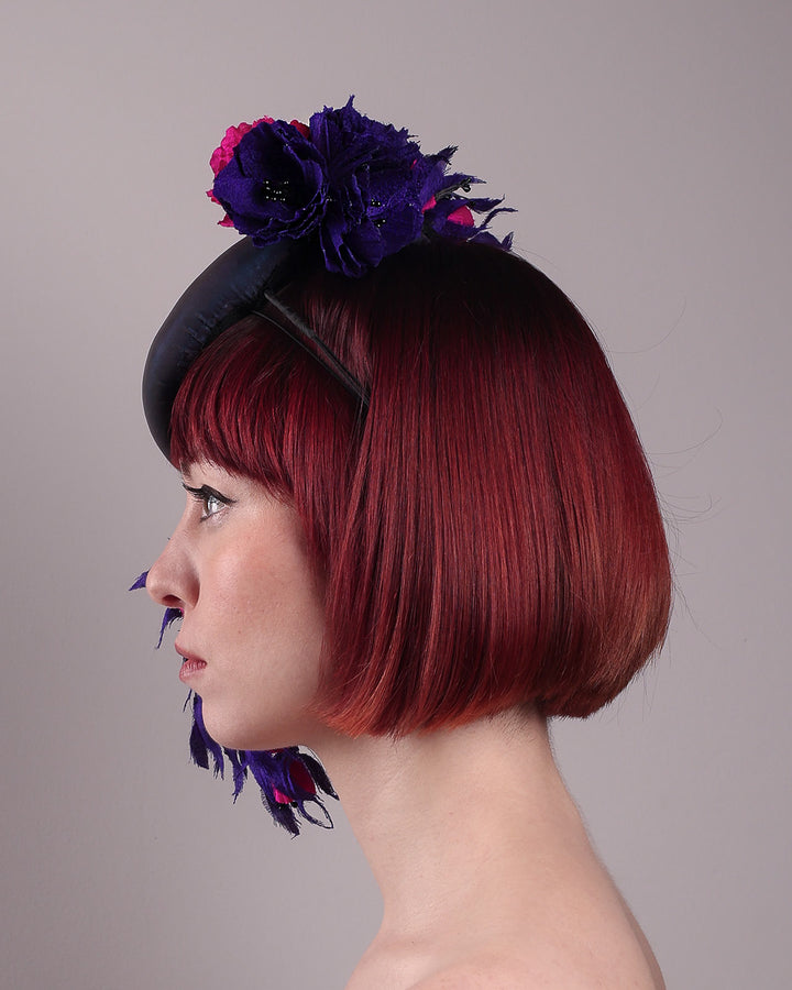 The model is showing the left side view of a pillbox fascinator with an ombré finish that transitions from deep midnight to luminous violet under varying light conditions, on her head. Adorned with handcrafted violet and fuchsia flowers, they cascade asymmetrically from the back to the right side of the face, extending to under the chin. The pillbox is fitted with a sleek headband and an elastic. Designed by Merve Bayindir.