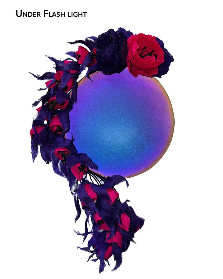 The still life picture under flash light, showing the colour of a pillbox fascinator with an ombré finish that transitions from deep midnight to luminous violet under varying light conditions. Adorned with handcrafted violet and fuchsia flowers, they cascade asymmetrically from the back to the right side of the face, extending to under the chin. The pillbox is fitted with a sleek headband and an elastic. Designed by Merve Bayindir.