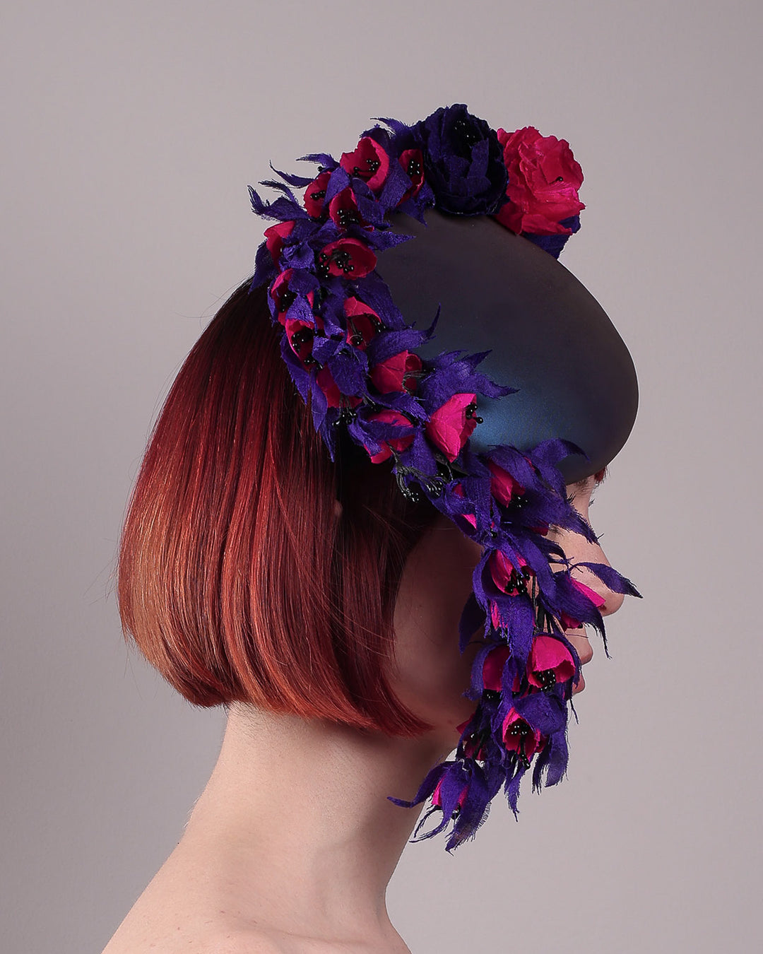 The model is showing the right side view of a pillbox fascinator with an ombré finish that transitions from deep midnight to luminous violet under varying light conditions, on her head. Adorned with handcrafted violet and fuchsia flowers, they cascade asymmetrically from the back to the right side of the face, extending to under the chin. The pillbox is fitted with a sleek headband and an elastic. Designed by Merve Bayindir.