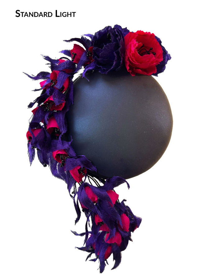 The still life picture standard flash light, showing the colour of a pillbox fascinator with an ombré finish that transitions from deep midnight to luminous violet under varying light conditions. Adorned with handcrafted violet and fuchsia flowers, they cascade asymmetrically from the back to the right side of the face, extending to under the chin. The pillbox is fitted with a sleek headband and an elastic. Designed by Merve Bayindir.