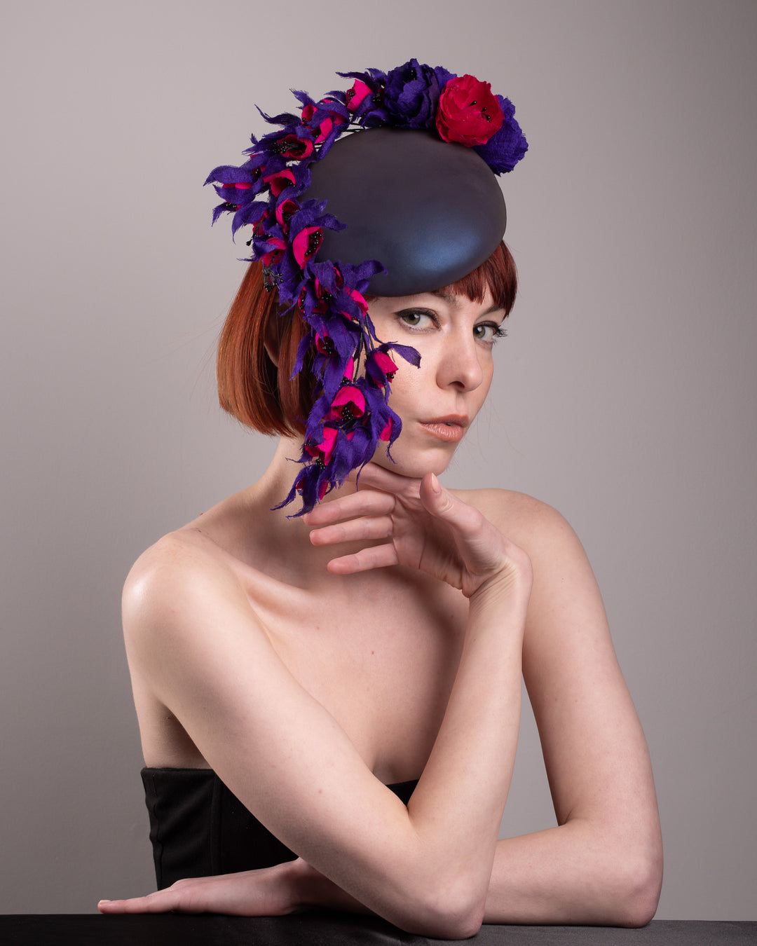 The model is showcasing a right front view of a pillbox fascinator with an ombré finish that transitions from deep midnight to luminous violet under varying light conditions. Adorned with handcrafted violet and fuchsia flowers, they cascade asymmetrically from the back to the right side of the face, extending to under the chin. The pillbox is fitted with a sleek headband and an elastic. Designed by Merve Bayindir.