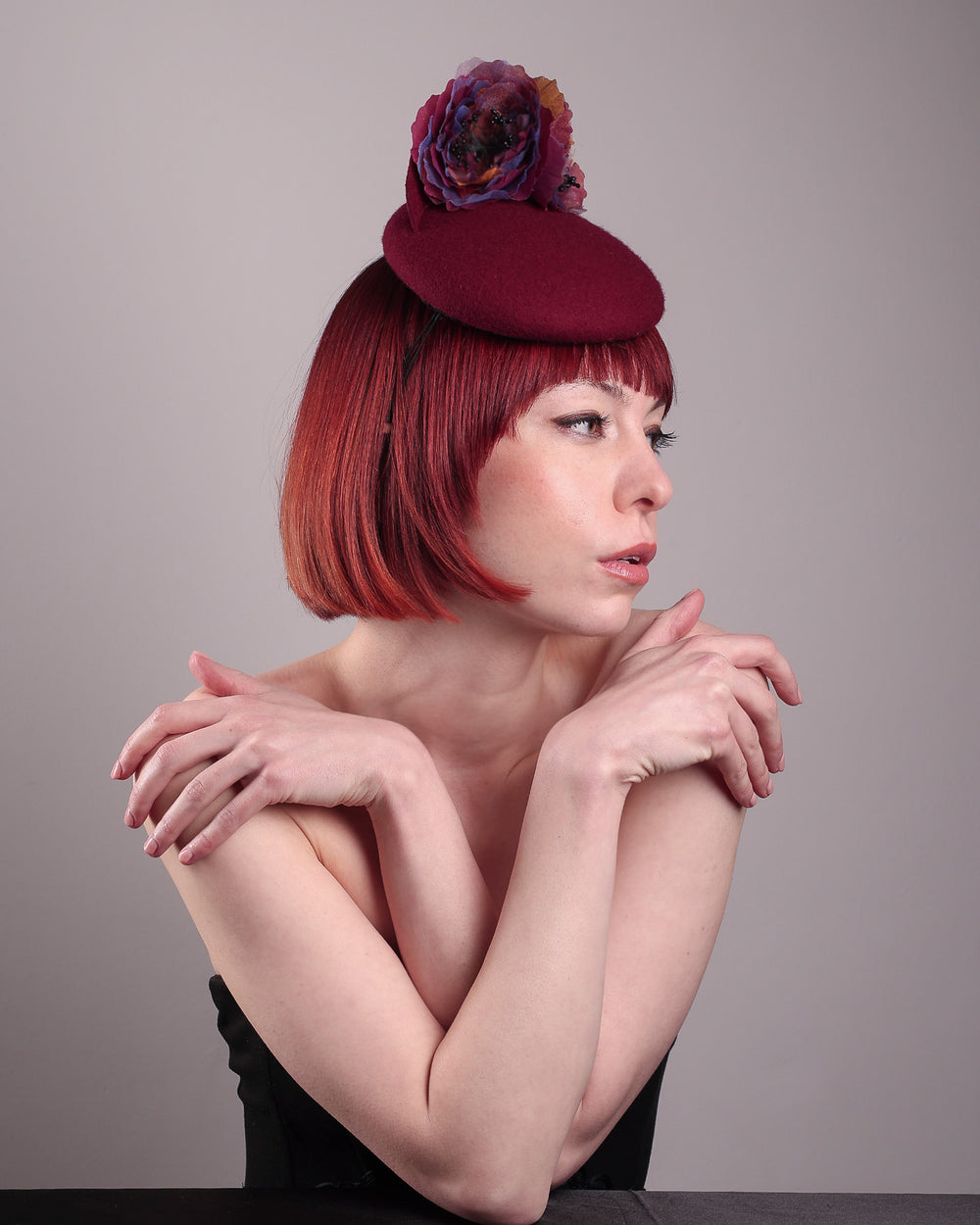 Model wearing and showcasing the right side of a burgundy felt pillbox fascinator with two handcrafted layered flowers in plum and bronze shades, featuring black bead centres, designed by Merve Bayindir