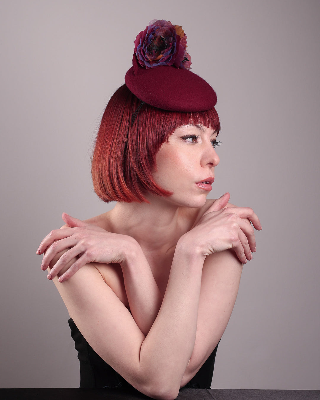 Model wearing and showcasing the right side of a burgundy felt pillbox fascinator with two handcrafted layered flowers in plum and bronze shades, featuring black bead centres, designed by Merve Bayindir