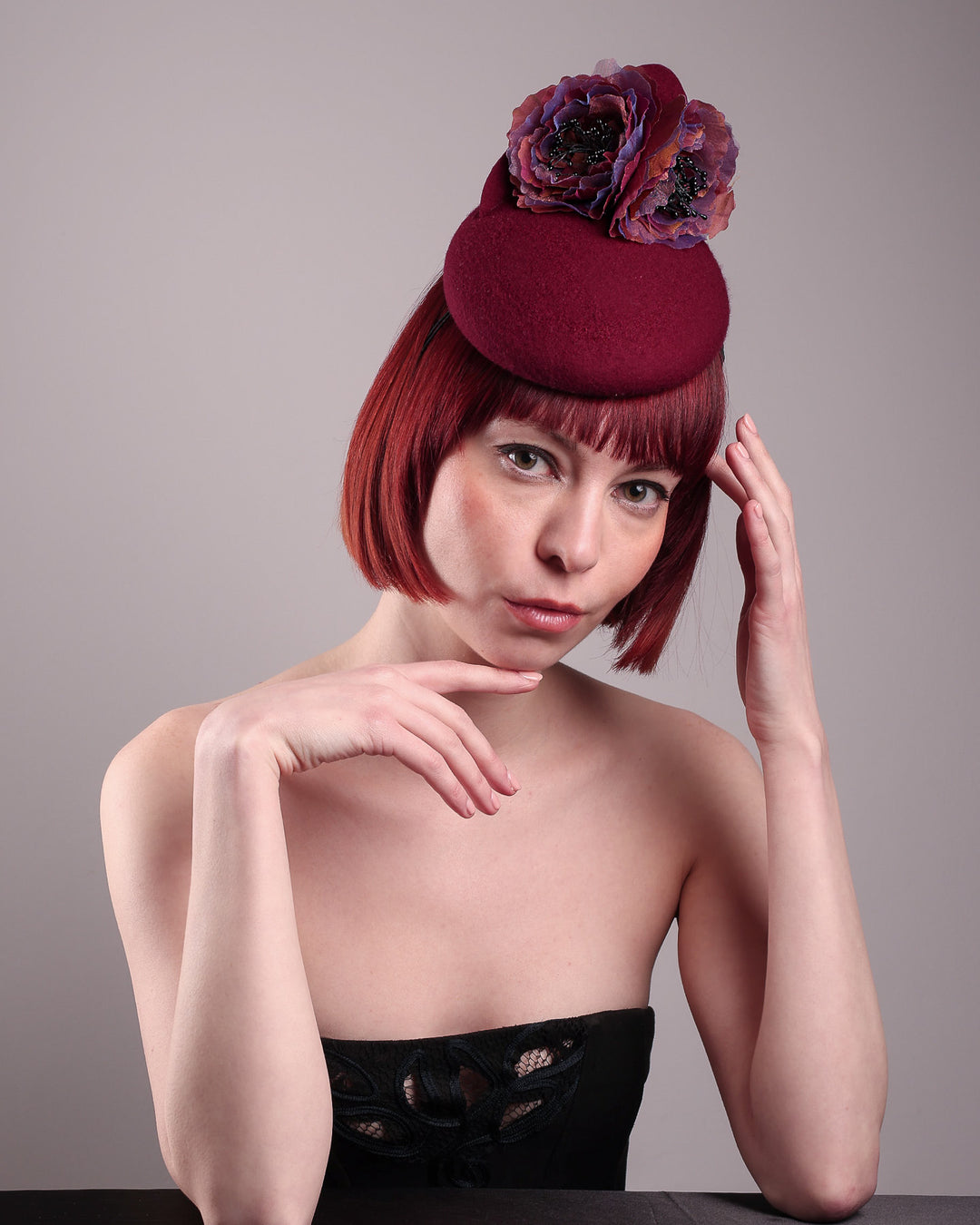 Model wearing and showcasing the front top angle of a burgundy felt pillbox fascinator with two handcrafted layered flowers in plum and bronze shades, featuring black bead centres, designed by Merve Bayindir