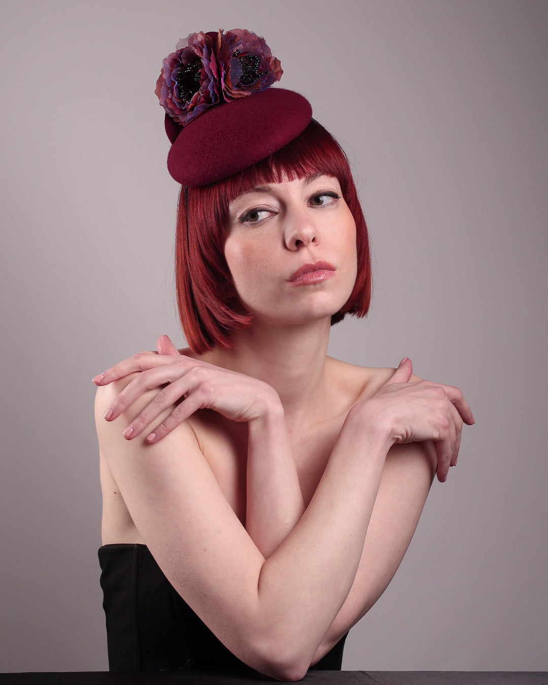 Model wearing and showcasing the front angle of a burgundy felt pillbox fascinator with two handcrafted layered flowers in plum and bronze shades, featuring black bead centres, designed by Merve Bayindir