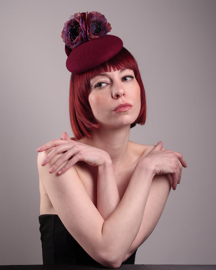 Model wearing and showcasing the front angle of a burgundy felt pillbox fascinator with two handcrafted layered flowers in plum and bronze shades, featuring black bead centres, designed by Merve Bayindir