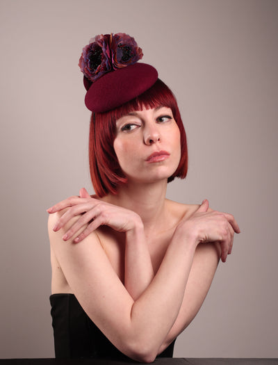 Model wearing and showcasing the front angle of a burgundy felt pillbox fascinator with two handcrafted layered flowers in plum and bronze shades, featuring black bead centres, designed by Merve Bayindir