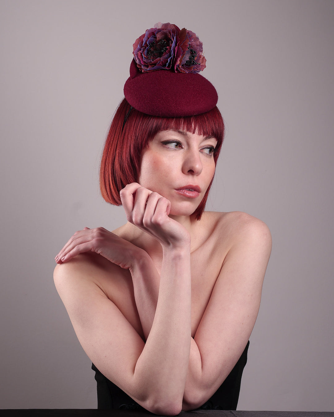 Model wearing and showcasing the front right angle of a burgundy felt pillbox fascinator with two handcrafted layered flowers in plum and bronze shades, featuring black bead centres, designed by Merve Bayindir