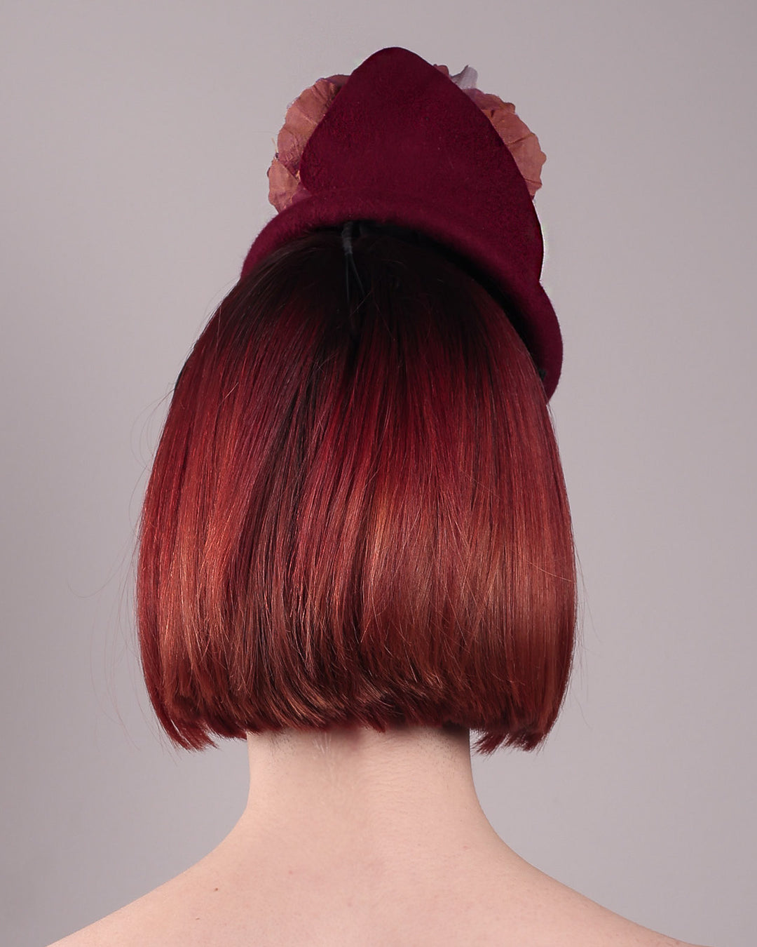 Model wearing and showcasing the back close up of a burgundy felt pillbox fascinator with two handcrafted layered flowers in plum and bronze shades, featuring black bead centres, designed by Merve Bayindir.