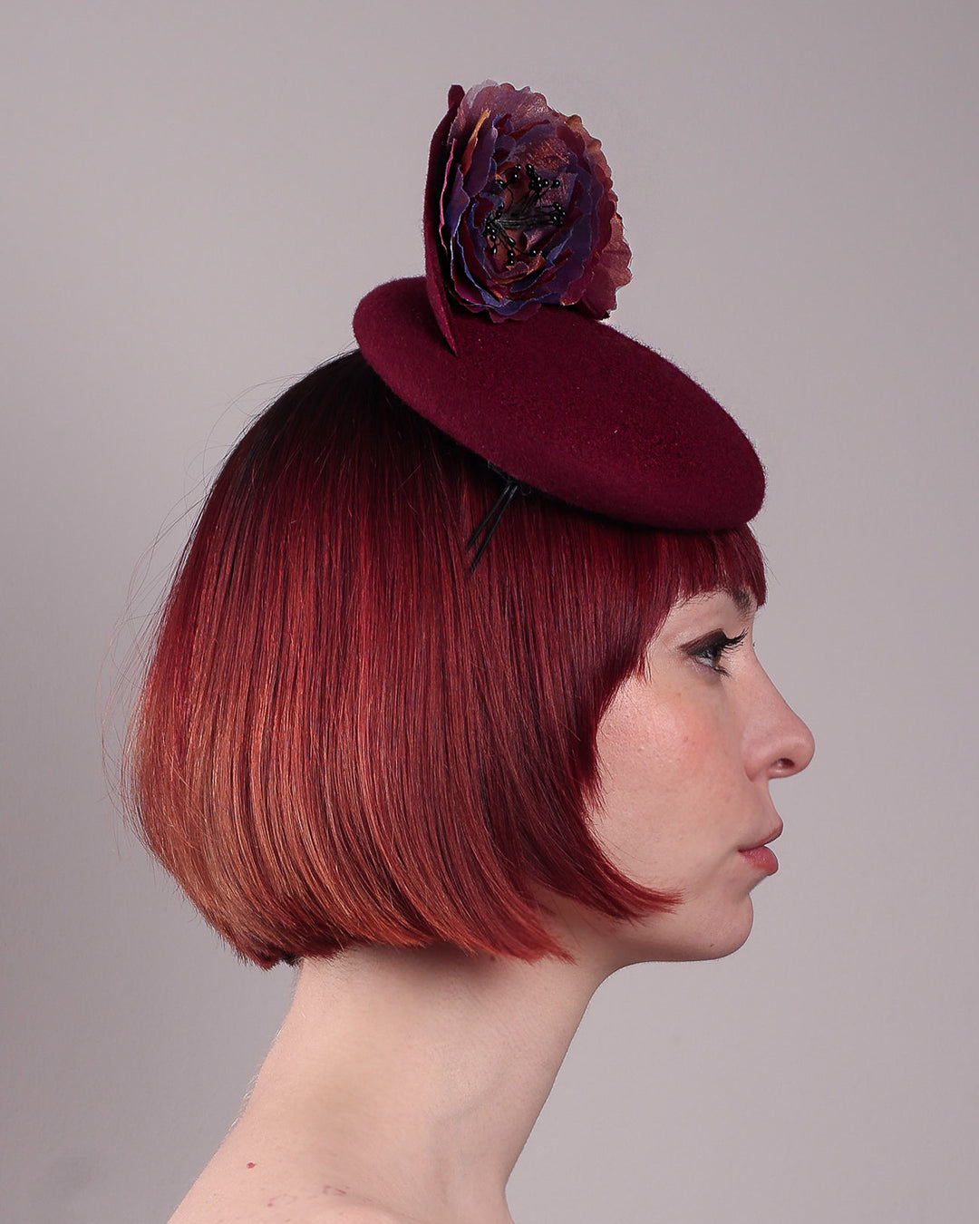 Model wearing and showcasing the right close up of a burgundy felt pillbox fascinator with two handcrafted layered flowers in plum and bronze shades, featuring black bead centres, designed by Merve Bayindir.