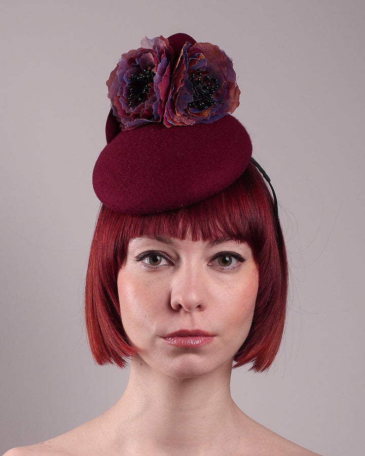 Model wearing and showcasing the front close up of a burgundy felt pillbox fascinator with two handcrafted layered flowers in plum and bronze shades, featuring black bead centres, designed by Merve Bayindir.