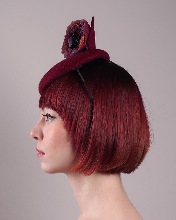 Model wearing and showcasing the left close up of a burgundy felt pillbox fascinator with two handcrafted layered flowers in plum and bronze shades, featuring black bead centres, designed by Merve Bayindir.