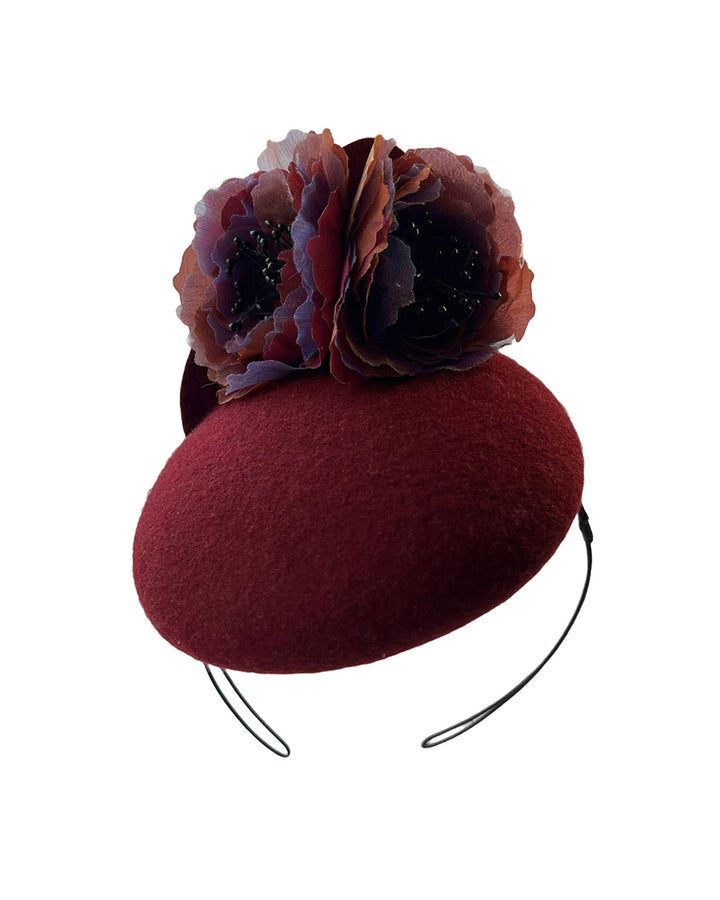 Still life photo of a burgundy felt pillbox fascinator with two handcrafted layered flowers in plum and bronze shades, featuring black bead centres, designed by Merve Bayindir.