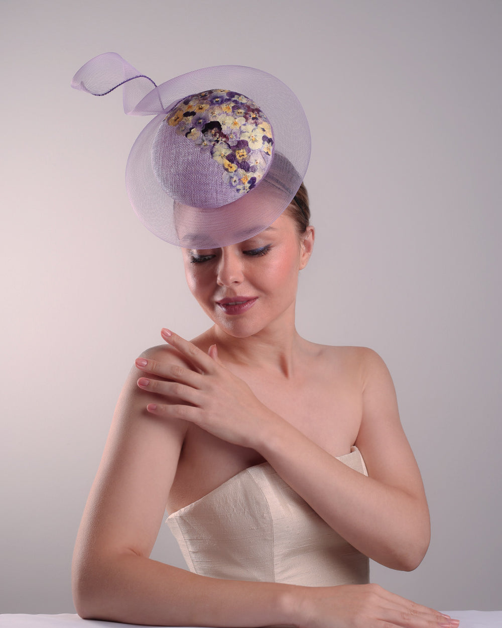 Model is showcasing the lilac double button fascinator from the left front side, worn to the right centre of the head. Decorated with pressed natural flowers on the left top side of the hat creating a complax motive. The button shape cirlcled with crinoline ending with a swirling edge at the top right side.