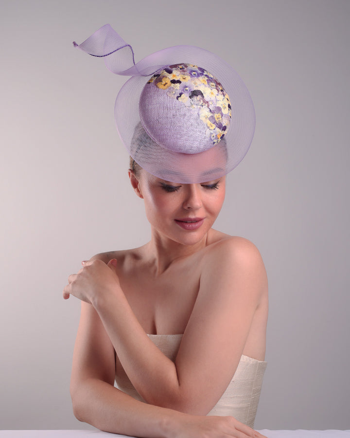 Model is showcasing the right front side of a lilac double button fascinator, worn to the right centre of the head. Decorated with pressed natural flowers on the left top side of the hat creating a complax motive. The button shape cirlcled with crinoline ending with a swirling edge at the top right side.