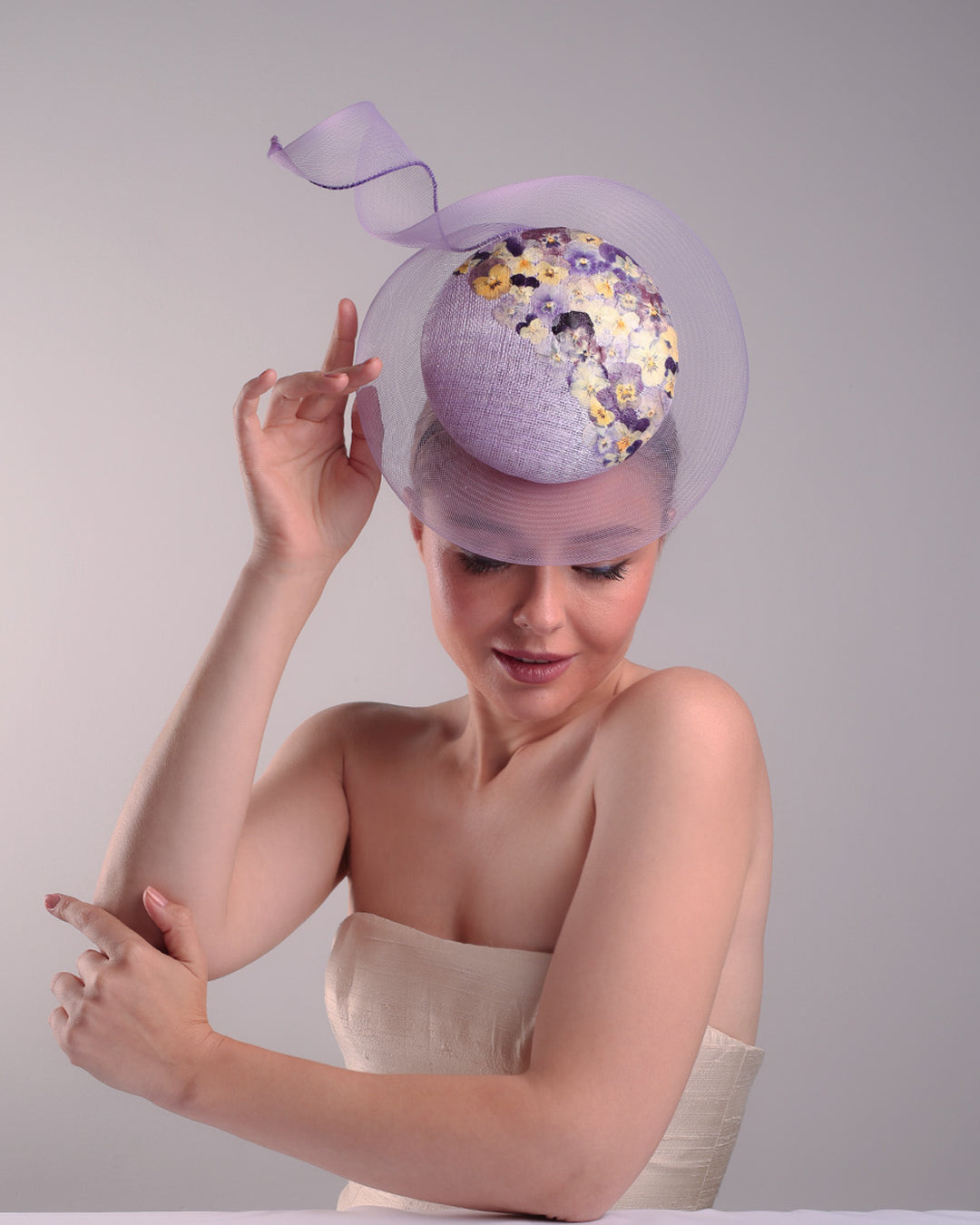 Model is showcasing the front of a lilac double button fascinator, worn to the right centre of the head. Decorated with pressed natural flowers on the left top side of the hat creating a complax motive. The button shape cirlcled with crinoline ending with a swirling edge at the top right side.