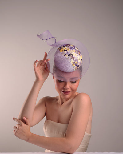 Model is showcasing the front of a lilac double button fascinator, worn to the right centre of the head. Decorated with pressed natural flowers on the left top side of the hat creating a complax motive. The button shape cirlcled with crinoline ending with a swirling edge at the top right side.