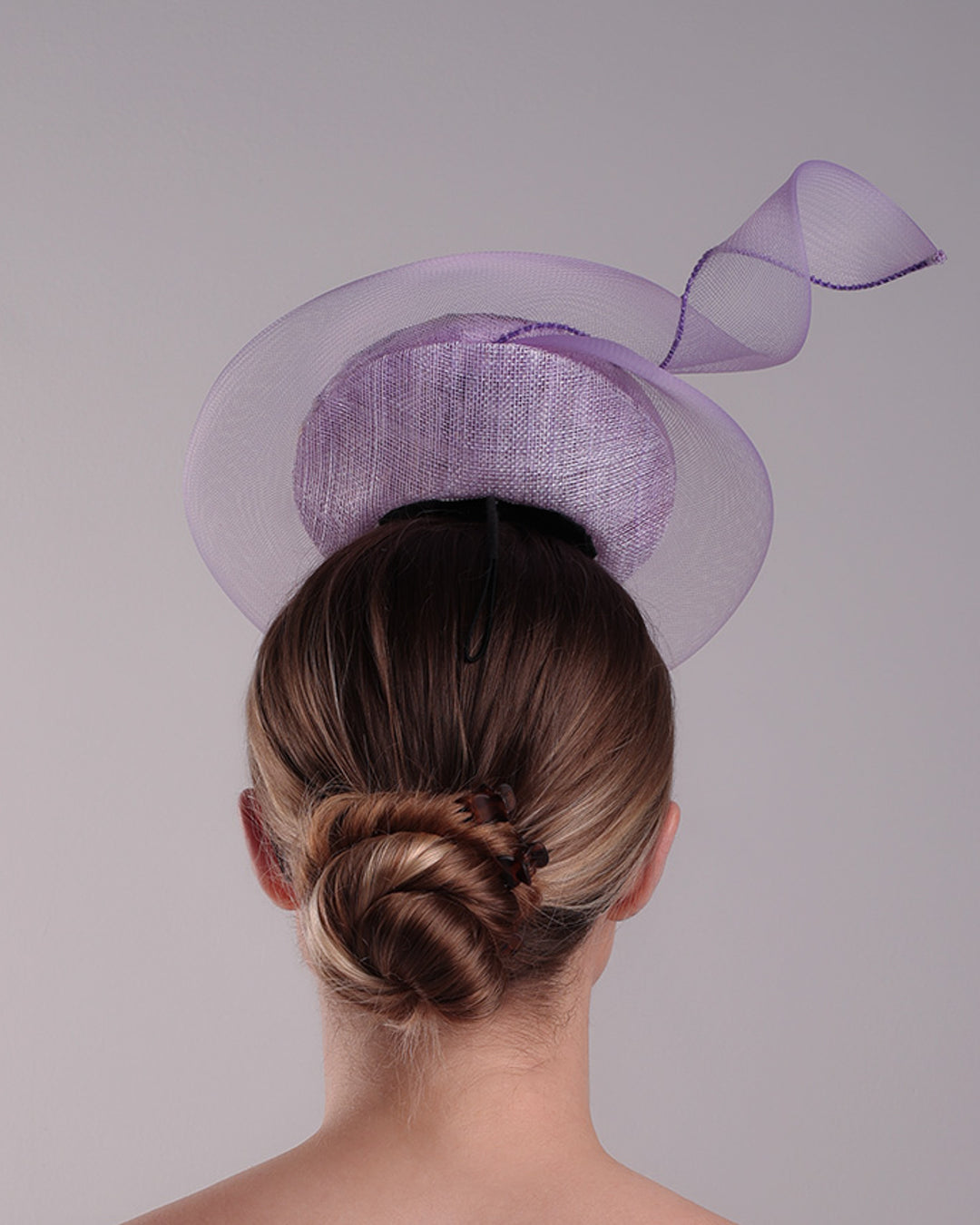 Model is showcasing the back of a lilac double button fascinator, worn to the right centre of the head. Decorated with pressed natural flowers on the left top side of the hat creating a complax motive. The button shape cirlcled with crinoline ending with a swirling edge at the top right side.