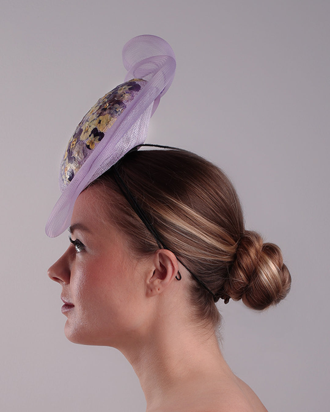 Model is showcasing the left side of a lilac double button fascinator, worn to the right centre of the head. Decorated with pressed natural flowers on the left top side of the hat creating a complax motive. The button shape cirlcled with crinoline ending with a swirling edge at the top right side.