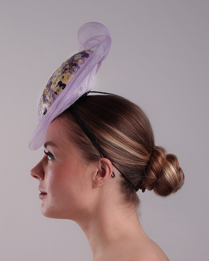Model is showcasing the left side of a lilac double button fascinator, worn to the right centre of the head. Decorated with pressed natural flowers on the left top side of the hat creating a complax motive. The button shape cirlcled with crinoline ending with a swirling edge at the top right side.