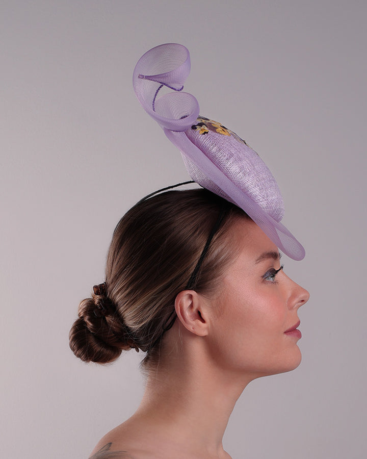 Model is showcasing the right side of a lilac double button fascinator, worn to the right centre of the head. Decorated with pressed natural flowers on the left top side of the hat creating a complax motive. The button shape cirlcled with crinoline ending with a swirling edge at the top right side.